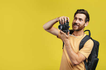 Positive middle aged man with backpack taking picture on professional DSLR camera over yellow...