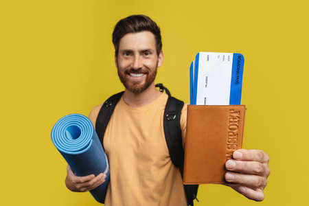 Excited man with backpack showing international passport and plane tickets on yellow studio...