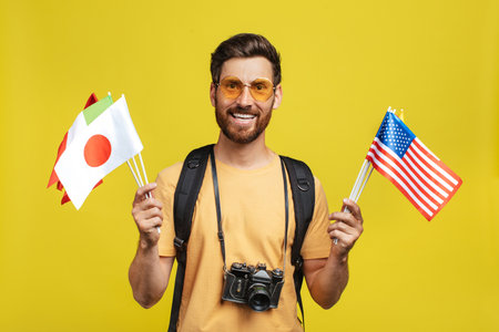 Travelling abroad. Male tourist holding international flags, standing with backpack and camera over...