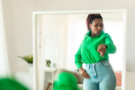 Playful african american woman dancing near mirror and smiling at her reflection, having fun in...
