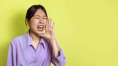 Japanese Woman Shouting Loudly Holding Hand Near Mouth, Yellow Backgroundの写真素材