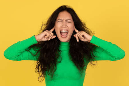 Woman Shouting With Eyes Closed Covering Ears Over Yellow Background