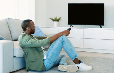 African american man watching television, pointing remote control at empty screen, mockupの写真素材
