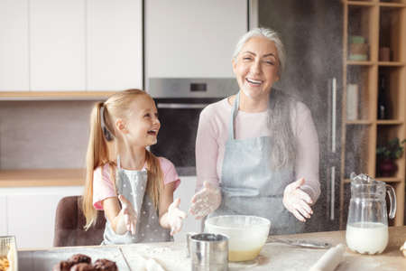 Cheerful little girl and senior grandma in aprons make dough, have fun with flour in cozy kitchen interiorの写真素材