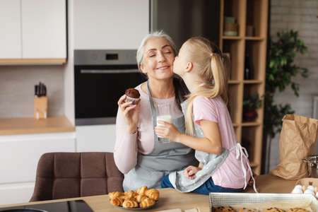 Cheerful european little granddaughter kisses her grandmother on cheek enjoy cookies with milk at...