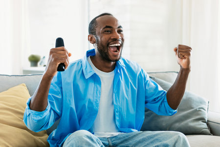 Overjoyed black man watching sport on TV at home, celebrating success, raising fists up and...