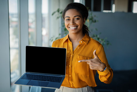 Happy african american businesswoman pointing at laptop computer with blank screen, standing in office, mockupの写真素材