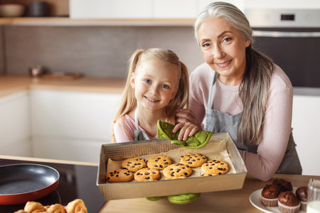 Smiling little girl and senior female in aprons show homemade fresh cookies in kitchen interiorの写真素材