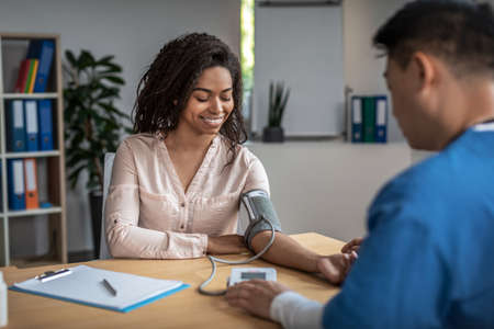 Adult asian man doctor measures pressure of smiling young black female patient in clinic office...