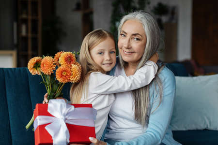 Cheerful european small girl hugging old woman with gift box and bouquet of flowers in living room...