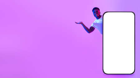 Happy young black man offering blank space from behind big cellphone with empty white screen in neon...