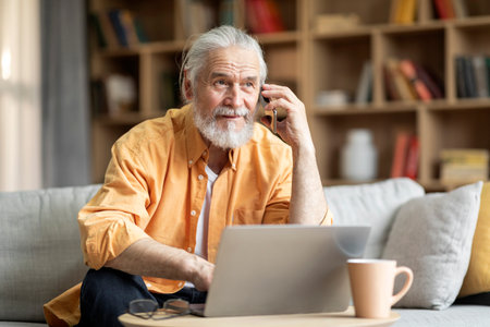 Cheerful elderly man using laptop, having phone conversationの写真素材