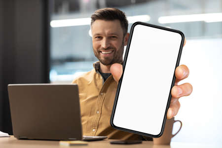 Business Offer. Smiling Millennial Man Demonstrating Big Blank Smartphone In Office