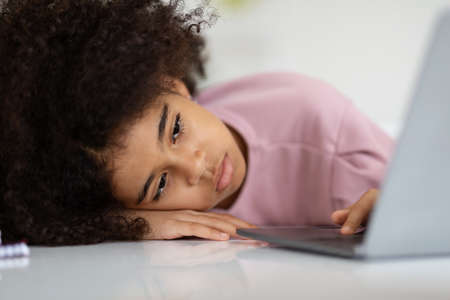 Closeup of bored black girl lying on desk with laptopの写真素材