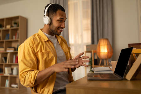 Happy young black man using laptop, wearing headphones, having video chat at home, copy space
