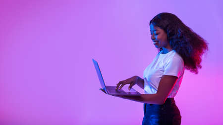 Happy young black lady using modern laptop, studying remotely or having business meeting in neon light, panoramaの写真素材