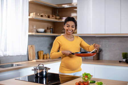 Happy young pregnant african american female with big belly holding frying pan and cooking food