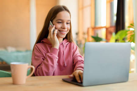 Portrait of happy teen girl talking on cellphone and typing on laptop keyboard, sitting at desk, talking with friendsの写真素材