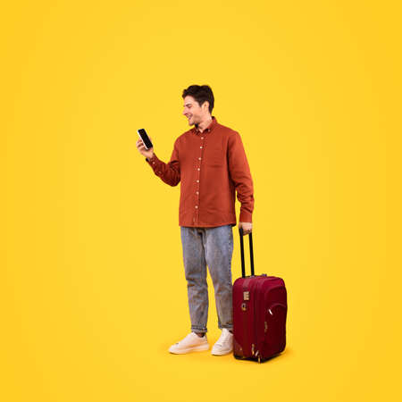 Traveler Man With Suitcase Using Cellphone Texting On Yellow Backgroundの写真素材