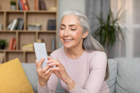 Smiling caucasian old gray-haired female typing on smartphone, have online call in living room