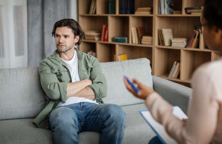 Dissatisfied young caucasian guy client ignores doctor at meeting with black lady psychologist in officeの写真素材