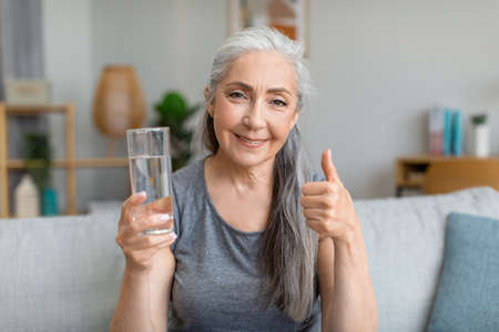 Happy pretty caucasian senior gray-haired female holds glass of water and shows thumb upの写真素材