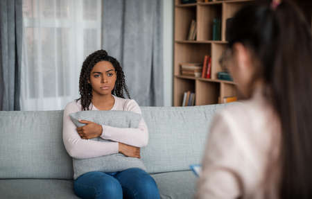 Consultation with psychologist. Despaired young african american female looking at caucasian doctor in officeの写真素材