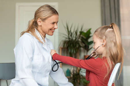 Happy little girl visiting pediatrician and playing with doctor, listening docs heart beating with...