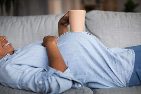 Unrecognizable Pregnant Black Female Holding Cup Lying On Sofa Indoorの写真素材