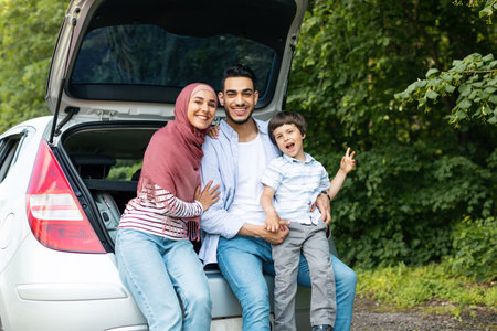 Glad smiling young middle eastern husband and wife in hijab holding baby, boy show peace sign with handの写真素材