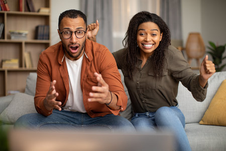 African American Spouses Shaking Fists In Joy Watching TV Indoorsの写真素材