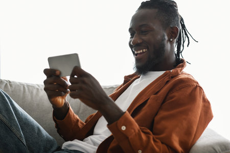 Excited african american male gamer playing games on smartphone while chilling on couch at homeの写真素材