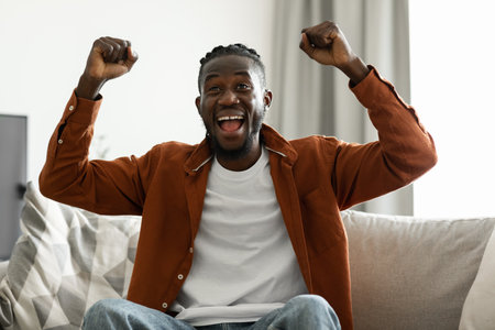Joyful black man cheering for sports team on television, gesturing YES and shouting, sitting on sofa at homeの写真素材