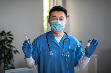 Glad adult japanese man doctor in protective mask and gloves shows syringe and vaccine in clinic interiorの写真素材