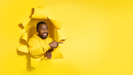 Happy african american man looking aside through hole in yellow paper and pointing aside with his...