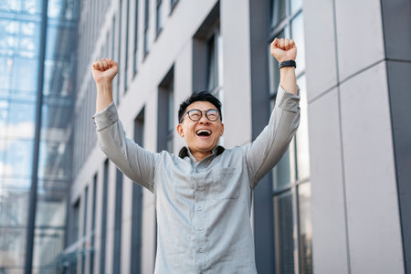 Business win. Overjoyed asian businessman shaking fists and celebrating success, standing outdoors, free spaceの写真素材