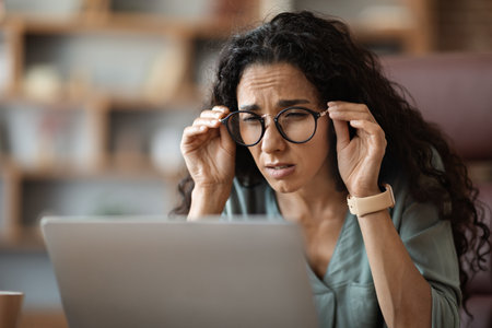 Woman sitting at desk in front of laptop, touching eyeglassesの写真素材