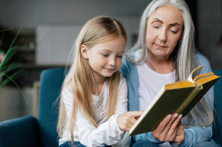 Serious caucasian small granddaughter and mature grandmother are reading book on sofa in room interiorの写真素材