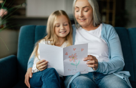 Cheerful caucasian small granddaughter and mature grandmother give drawing postcard and read on sofaの写真素材
