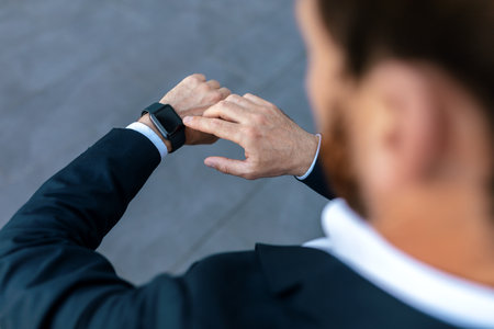 Young caucasian guy with beard in suit checks time on smartwatch with blank screen on street,...