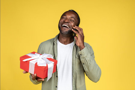Laughing mature black male in casual talking on smartphone and holding gift boxの写真素材