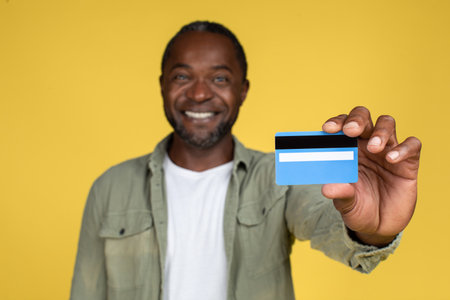 Glad cheerful middle aged black male in casual showing credit card, isolated on yellow background, studioの写真素材