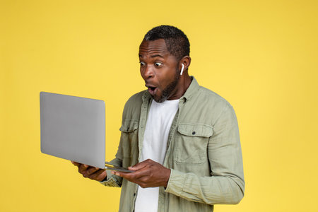 Surprised happy funny adult african american male in casual and headphones looking at laptop