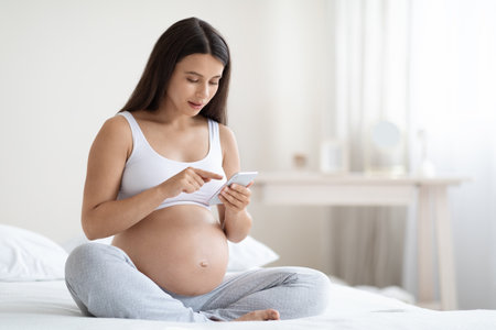 Peaceful expecting lady using cell phone, bedroom interiorの写真素材