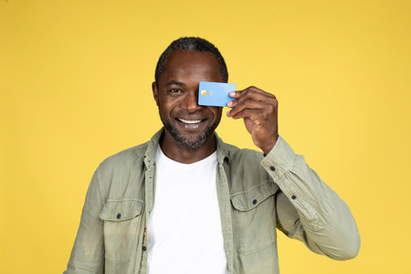 Funny happy adult black guy in casual puts credit card to eye, rejoices to great online shopping,...