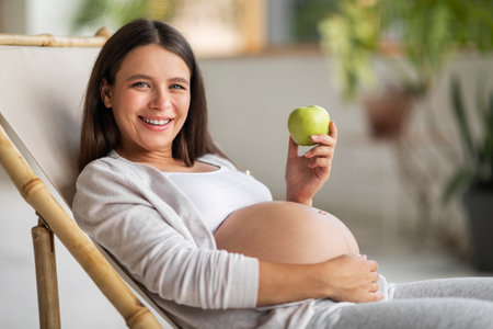 Healthy Snack. Beautiful pregnant woman sitting in chair and holding green appleの写真素材