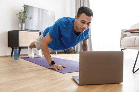 Handsome arab guy in sportswear doing push-ups, having online fitness class via laptop from home,...