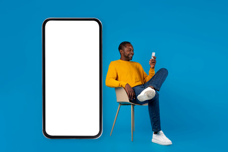 African american man sitting on chair by huge smartphone, mockupの写真素材