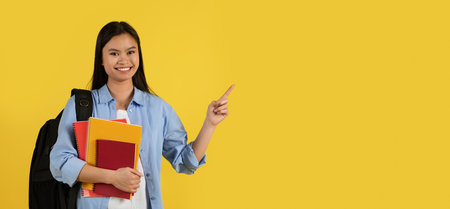 Smiling young chinese female student in casual with backpack and many books pointing finger at empty...
