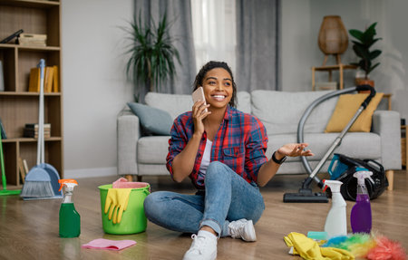 Happy young black woman in rubber gloves sit on floor with cleaning supplies, calling by phone in living roomの写真素材